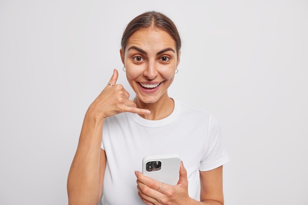 Young glad brunette woman makes phone gesture makes call me back sign holds smartphone smiles happily dressed in casual t shirt isolated over white wall