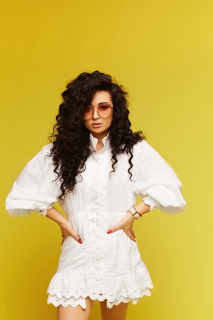 Young glamorous model woman wearing a white dress and trendy sunglasses posing over yellow background