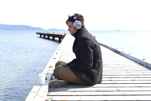 Young gloomy man listening to music sitting all alone on pier on a windy spring day