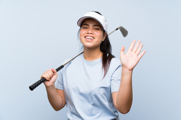 Ragazza asiatica del giovane giocatore di golf sopra la parete blu isolata che saluta con la mano con l'espressione felice