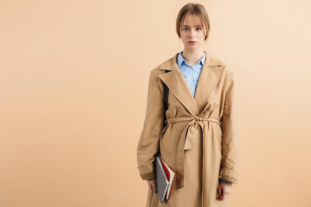 Young gorgeous lady in trench coat and blue shirt holding notepads in hand dreamily looking in camera over beige background isolated