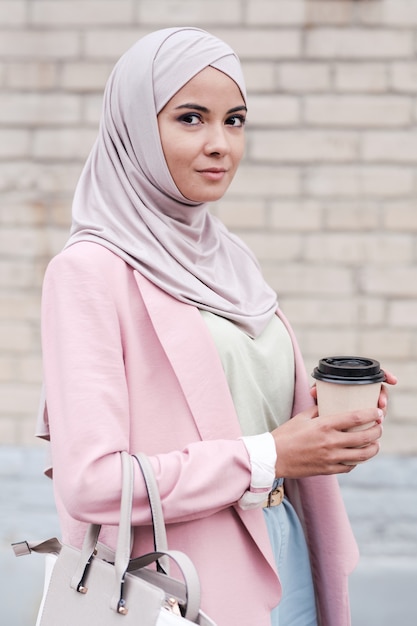 Молодая великолепная мусульманка в хиджабе, пуловере и розовом кардигане пьет кофе, стоя в городской среде