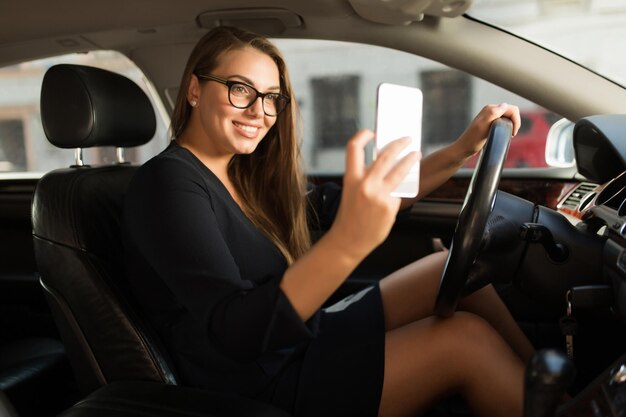 Young gorgeous smiling woman in black dress and eyeglasses happily taking photo on cellphone while sitting behind the wheel in car