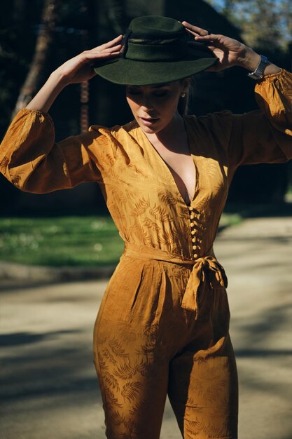 Young gorgeous woman in bright overalls wearing hat sensually posing on camera outdoor