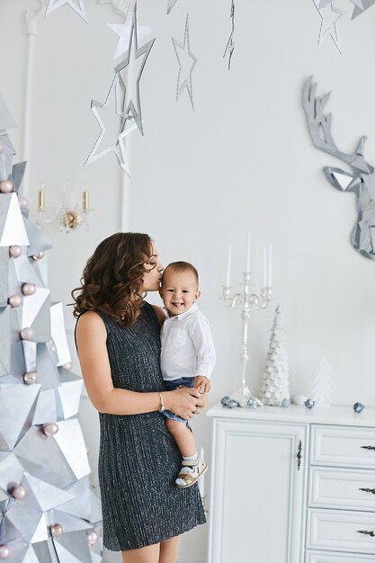 Young gorgeous woman in a fashionable dress holds in her hands her cute son at the interior decorated for the New year party