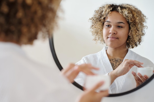 Foto giovane donna splendida che si guarda allo specchio mentre si applica la crema sul viso