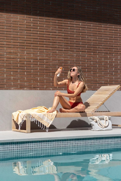 Young gorgeous woman sitting on a sun lounger smiling while putting face sun cream on in the swimming pool