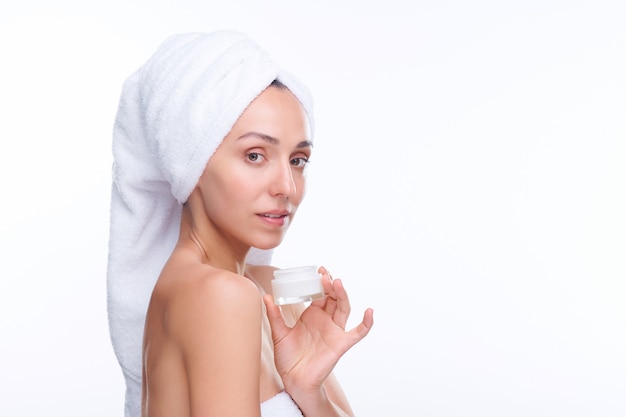 Young gorgeous woman with white towel on head holding jar of moisturizing facial cream while showing it to you in isolation