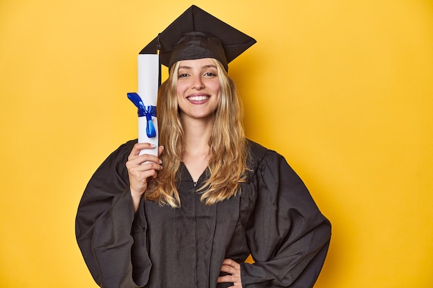 Photo young graduate woman in cap and gown holding a diploma on a yellow studio background