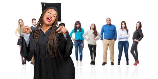 Young graduated black woman wearing braids pointing to the side, smiling surprised presenting something, natural and casual