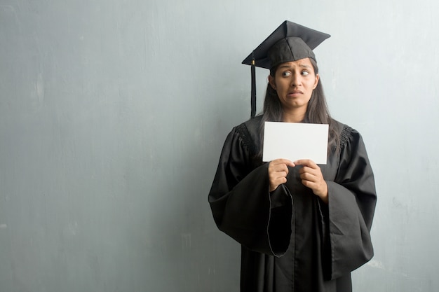 Young graduated indian woman against a wall very scared and afraid, desperate for something, cries of suffering and open eyes, concept of madness. Holding a placard.