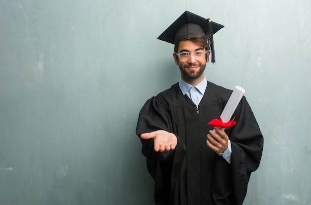 Young graduated man against a grunge wall with a copy space holding something with hands, showing a product, smiling and cheerful, offering an imaginary object