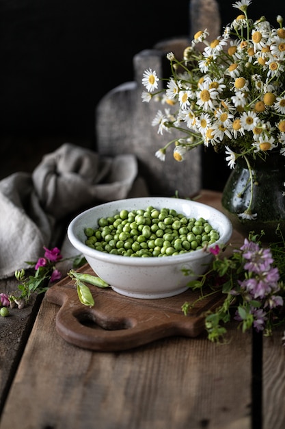 Young green peas in white bowl on wooden. Pea flowers and daisy flowers on the table.