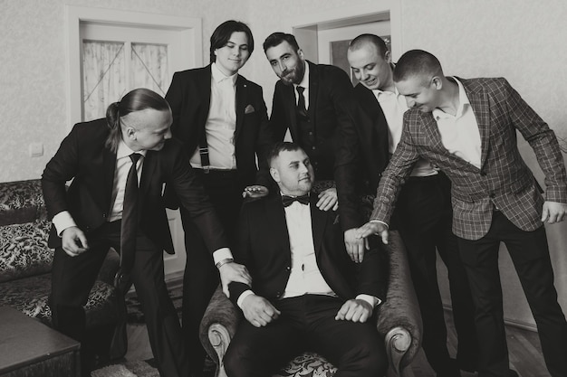A young groom and his funny friends pose for the camera A group of young people hug the groom and cheerfully congratulate him Happy friends Friends in the room Wedding day Black and white photo