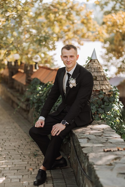 The young groom sits on the fence looking into the lens an adult man in a black suit and white shirt with a tie