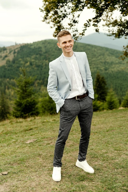 A young groom with a blue jacket stands near a tree against the backdrop of mountains