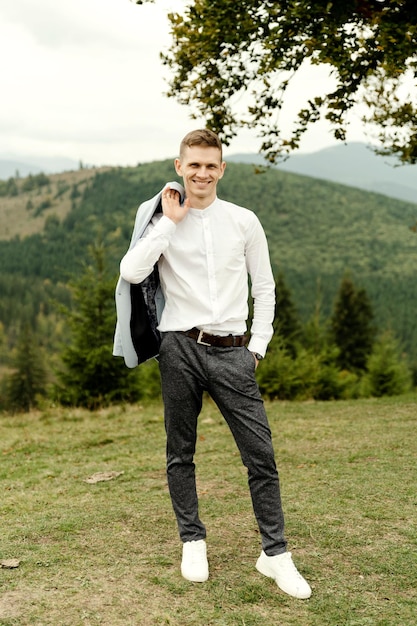 A young groom with a blue jacket stands near a tree against the backdrop of mountains