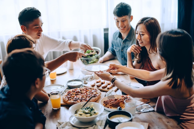 A young group of joyful friends having fun enjoying together passing and sharing food across dining table at party