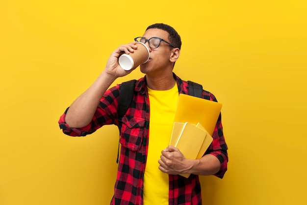 Молодой парень африканский студент в очках с рюкзаком пьет кофе на желтом фоне