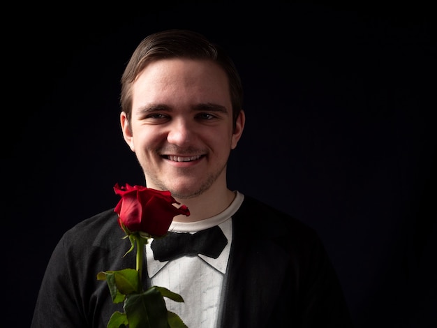 Il giovane ragazzo in un vestito nero della maglietta tiene una rosa rossa nelle sue mani e sorride sul nero