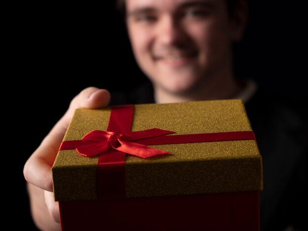 Giovane ragazzo in abito t-shirt nero allunga la mano con un regalo rosso e oro su fondo nero