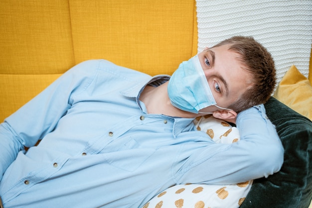 A young guy of caucasian ethnicity in a shirt and a protective mask sits on the couch alone at home ...