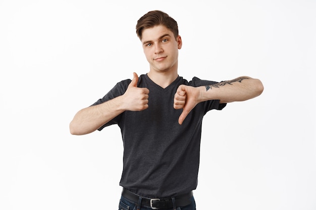 Young guy, college student show thumbs up thumb down, average rate, making decision, like or dislike, standing on white