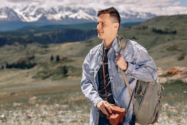 A young guy in a denim jacket with a backpack and a camera in a case stands on a green grassy lawn and looks away. Search for a suitable angle for a natural shot.