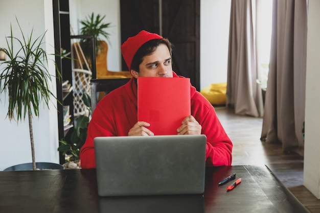 Young guy developer in red clothes with computer and note at home office