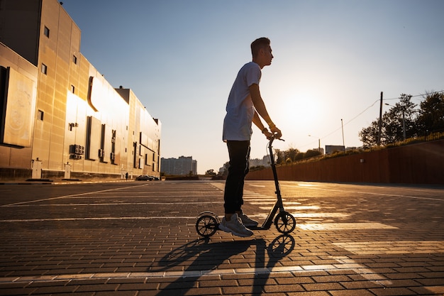 Young guy dressed in jeans and t-shirt is riding a scooter on the square paved with tiles near the building on the sunny day .