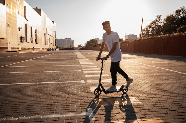 Young guy dressed in jeans and t-shirt is riding a scooter on the square paved with tiles near the building on the sunny day .