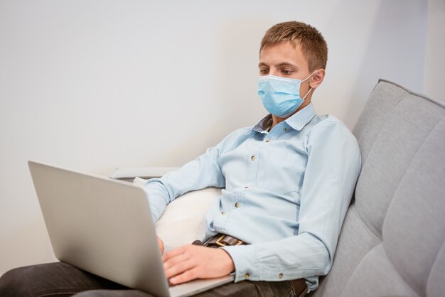 A young guy of european appearance in a light shirt sits in a protective medical mask and works on t...