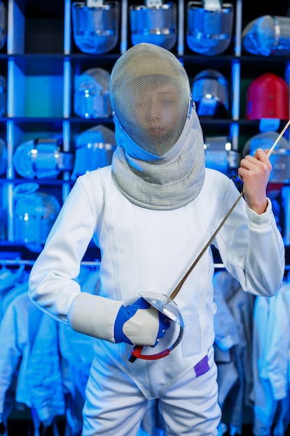 Young guy in a fencing suit with a sword in his hand, on a blue background, neon light. The athlete trains. Sports, youth, healthy lifestyle. Selective focus