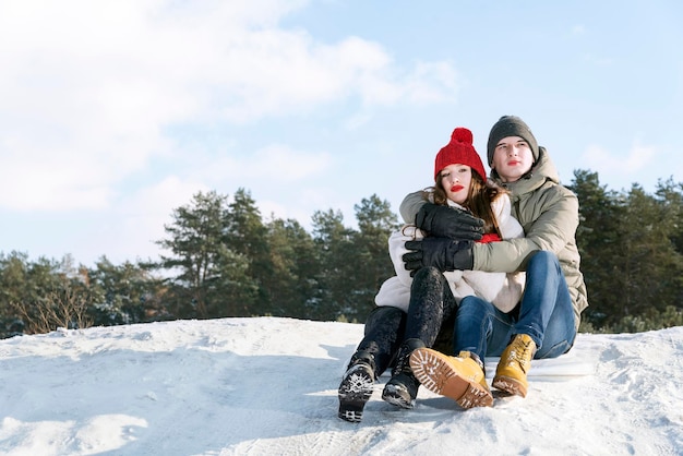 Young guy and a girl sit on the snow against the background of the forest and the blue sky. Couple outdoors. Copy space.