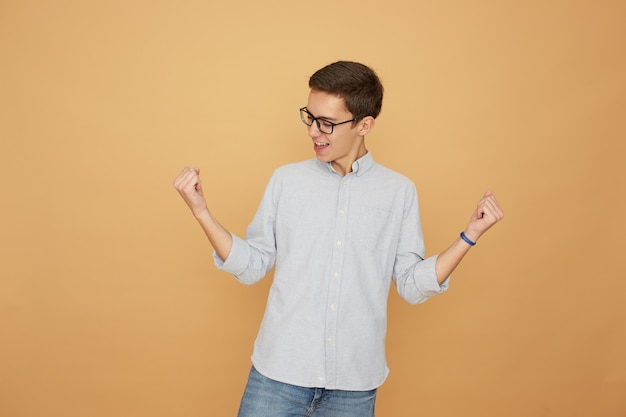 Young guy in glasses dressed in light blue shirt and jeans stands joyful on the beige background in the studio .