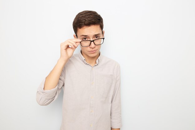 Young guy in glasses dressed in light shirt stands with his hand on his glasses on the white background in the studio .