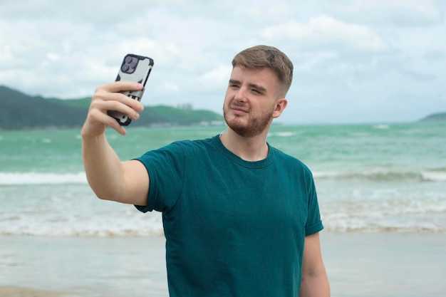 Young guy handsome happy man at sea is taking picture of hisself selfie at camera of his phone using smartphone for social media at summer beach in tropical exotic country Blogger vlog