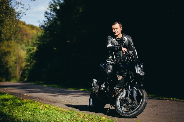 Фото Молодой парень в черной кожаной куртке сидит на спортивном мотоцикле на фоне красивой лесной дороги