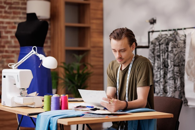 A young guy is a fashion designer He looks at the sketch while sitting at a table with a sewing machine