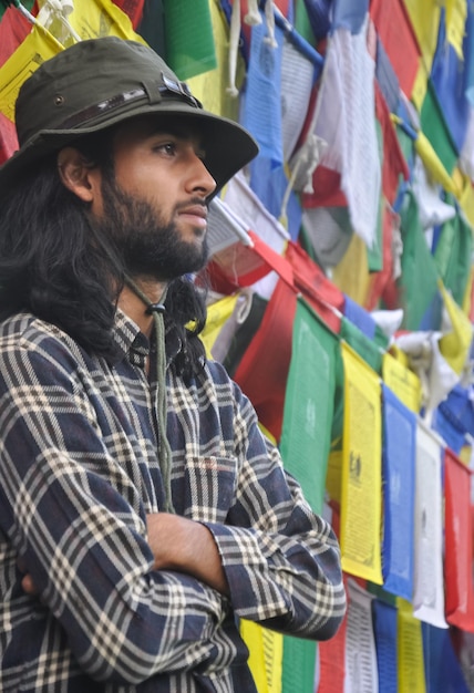 A young guy looking sideways posing with his arms crossed standing against buddhist prayer flags