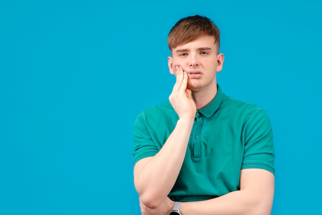 Young guy, a man suffering from toothache, holds his cheek with his hand. On a blue, monochrome background in the studio.