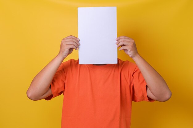 Young guy in orange tshirt hiding his face under blank white paper isolated yellow background