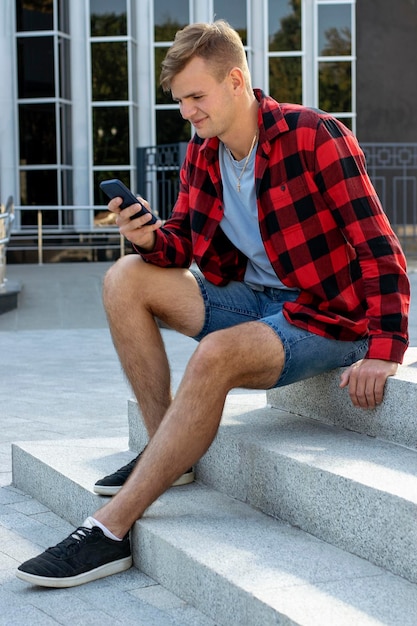 A young guy in a plaid shirt sits with a phone on the street