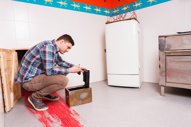 Young guy in plaid shirt squatting and opening the safe trying to get out of the kitchen with a bloody floor, escape the room game concept