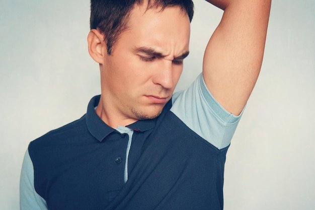 A young guy in a polo shirt sniffs his smelly armpit and makes\
a disgruntled expression on blue background negative emotion facial\
expression feeling