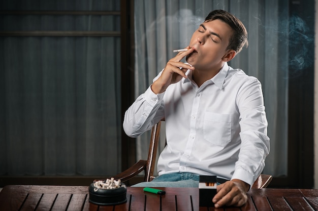 A young guy poses sitting at a table on which stands an ashtray full of cigarettes