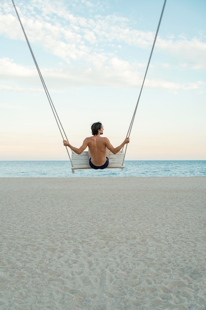 Young guy on rope swing on the beach on sea background Back view Vertical frame Rest on the sea