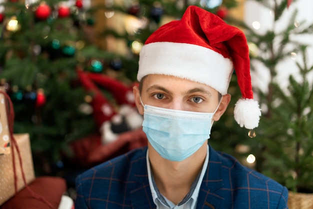 A young guy in a shirt and a santa claus hat on the background of a Christmas tree. Holidays during a pandemic