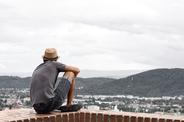 Photo a young guy sit on the edge of a tall building and looks into the distance towards the mountains