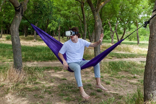 Photo a young guy sits in a purple hammock wearing virtual reality glasses 3d visualization and explores the space with his hands